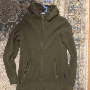 Olivgrön stickad kofta med huva - Säljer en olivgrön stickad kofta med dragkedja och huva. Koftan har långa ärmar och ribbade muddar vid ärmslut och nederkant. Perfekt för kyligare dagar.