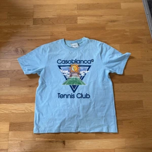 Ljusblå t-shirt från Casablanca - Säljer en ljusblå t-shirt från Casablanca med ett coolt tryck av en sol och tennisbana på framsidan. Perfekt för en avslappnad stil med en touch av sportig elegans.