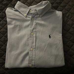 Blå skjorta från Ralph Lauren - Snygg ljus blå skjorta från Ralph Lauren med klassisk krage och knappar framtill. Skjortan har en broderad logotyp på bröstet och är långärmad. Perfekt för en stilren look. Skjortan är i bra skicka 7/10 och inga defekter. Är i storlek (XL Barn) vilket motsvarar S. 
