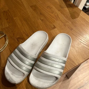 Vita sandaler från Adidas - Säljer ett par vita sandaler från Adidas med de klassiska tre ränderna i grått. Perfekta för sommarens varma dagar. Sandalerna har en enkel och stilren design.