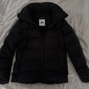 Svart dunjacka från J.Lindeberg - Säljer en stilren svart dunjacka från J.Lindeberg. Jackan har en praktisk huva och dragkedja framtill. Perfekt för kyliga dagar med sin vadderade design som håller dig varm. Två fickor med dragkedja på sidorna för säker förvaring.