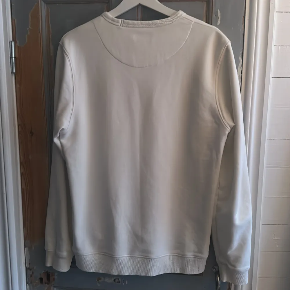 Säljer en stilren vit sweatshirt från Morris med rund hals och ett litet broderat emblem på bröstet. Tröjan har långa ärmar och ribbade muddar vid ärmslut och nederkant. Perfekt för en avslappnad look.. Neuletakit & Villapaidat.