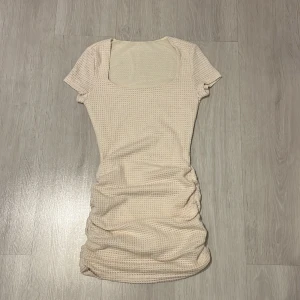 Beige stickad klänning - Säljer en söt beige stickad klänning med korta ärmar och en fyrkantig halsringning. Klänningen har en åtsittande passform med rynkade detaljer längs sidorna som ger en fin siluett. Perfekt för en stilren look!
