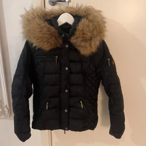 Svart dunjacka från Rockandblue med pälskrage - Säljer en snygg svart dunjacka från Rockandblue med en stor, mysig äkta päls på huvan. Jackan har dragkedja och knappar framtill samt flera praktiska fickor. Perfekt för kalla vinterdagar! 🖤