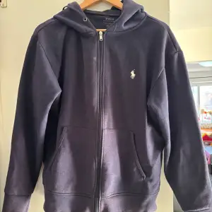 Säljer en stilren mörkblå hoodie från Polo Ralph Lauren i bra skick. Den har en dragkedja framtill och en klassisk logga på bröstet. Perfekt för både höst och vår, och passar till alla tillfällen. Superbekväm och snygg! 