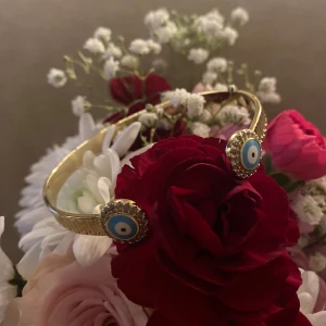 Guld färg rostfritt stål armband och halsband med ögonmotiv - Säljer ett snyggt guldarmband och matchande halsband med ögonmotiv. Armbandet har två blå och vita ögon med glittrande detaljer runtom. Halsbandet har ett hängsmycke med ett liknande öga och små rektangulära hängen. Perfekt för att ge en mystisk touch till din outfit!