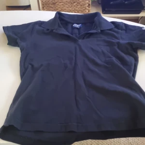 Svart pikétröja från James Harvest - Säljer en klassisk svart pikétröja från James Harvest Sportswear. Den har korta ärmar och en stilren krage, perfekt för både vardag och sportiga aktiviteter. Materialet känns bekvämt och hållbart. Passar bra till jeans eller kjol för en avslappnad look.