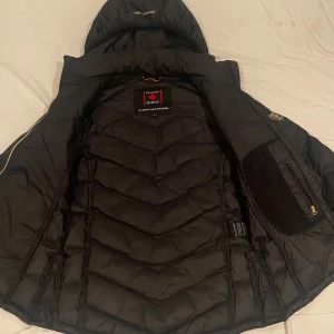 Svart dunjacka från Canada Snow - Säljer en snygg svart dunjacka från Canada Snow. Den har en design med guldiga dragkedjor och en huva för extra värme. Perfekt för kalla vinterdagar! Jackan har ett märke på ärmen med en guldfärgad lönnlöv. Passar både till vardags och lite finare tillfällen.