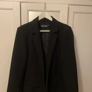 Svart kavaj från Pieces - Säljer en stilren svart kavaj från Pieces i storlek M. Perfekt för både vardag och fest. Kavajen har en klassisk design med långa ärmar och en snygg passform. Den är i mycket bra skick och redo att användas direkt. Passar perfekt till höst- och vårgarderoben!