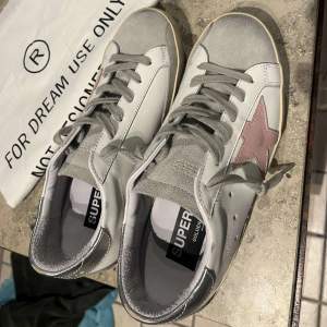 Säljer ett par snygga Golden Goose sneakers i vitt och grått med en rosa stjärna på sidan. Skorna har en cool vintage-look med silverdetaljer på hälen och grå snörning. Perfekta för en avslappnad men stilren look! Det är i storlek 38 och det är köpta på Grailed. Hör av er vid frågor!! 