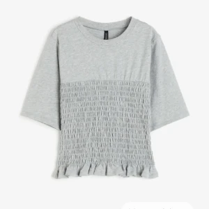 Grå topp med smockad midja - Säljer en söt grå topp med trekvartsärm och volangdetalj längst ner. Toppen har en ribbad struktur på mitten som ger en fin textur. Perfekt för vardagsbruk eller en festligare tillställning