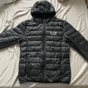 Svart pufferjacka från Emporio Armani - Säljer en snygg svart pufferjacka från Emporio Armani EA7. Jackan har en dragkedja framtill och en huva för extra skydd mot väder. Perfekt för kyliga dagar och har en stilren design med märkets logga på bröstet.