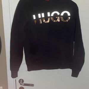 Svart tröja från Hugo - Säljer en snygg svart tröja från Hugo med ett coolt, glänsande logotryck på framsidan. Perfekt för både vardag och fest. Tröjan har långa ärmar och en bekväm passform. Passar perfekt till jeans eller chinos för en stilren look. Den är Xs men passar mer som S. Loggan flashar när man tar kort på den.