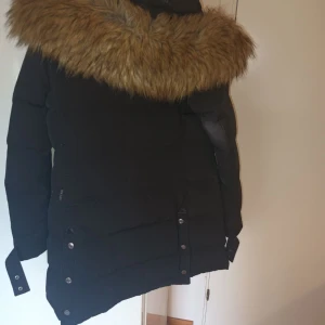 Svart vinterjacka från Hollies - Säljer en snygg svart vinterjacka från Hollies med stor pälskrage. Jackan har dragkedja och knappar framtill, samt långa ärmar. Perfekt för kalla vinterdagar! 🖤