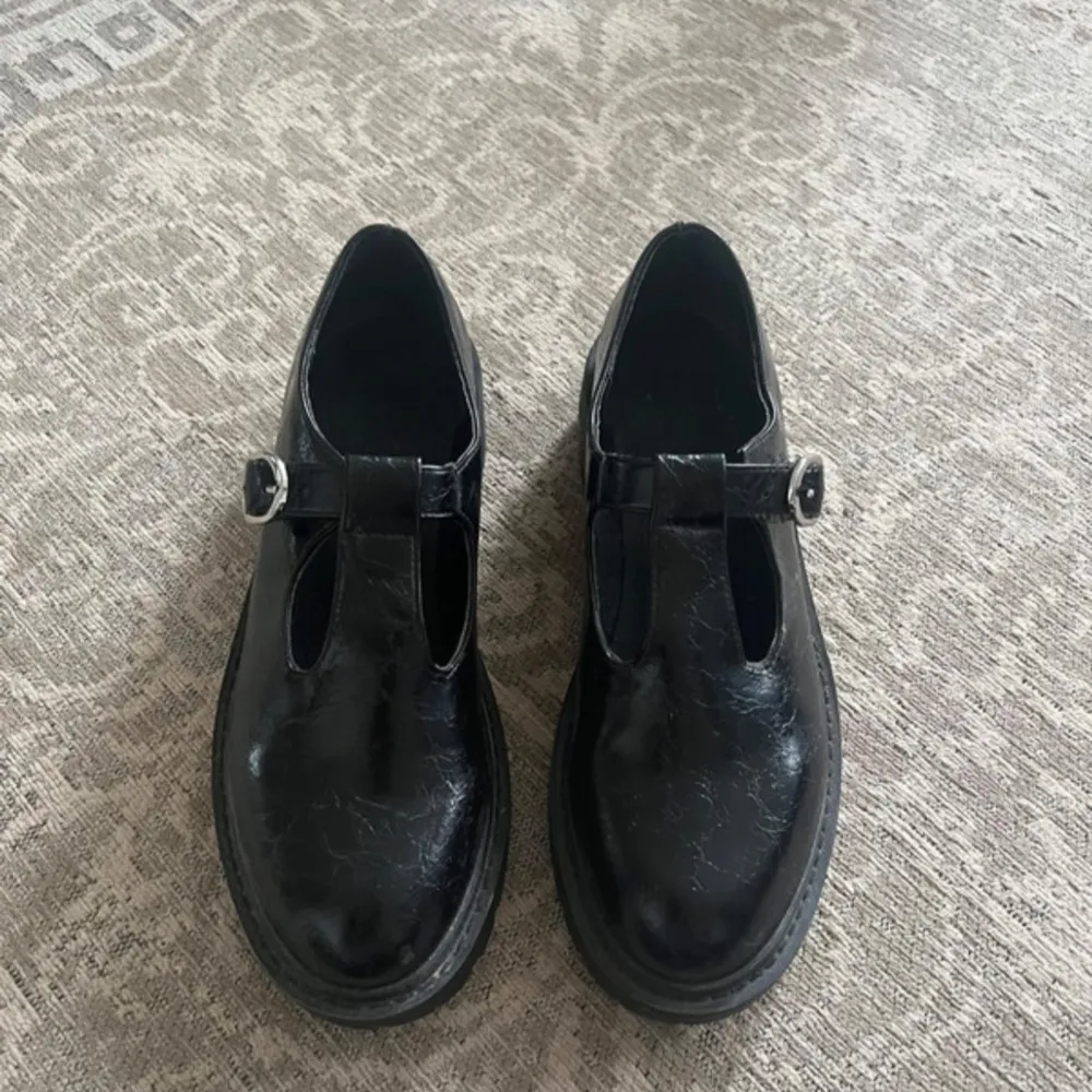 Säljer ett par snygga svarta loafers i skinn med ett coolt spänne över foten. De har en robust sula som ger bra grepp och en stilren design som passar perfekt till både vardag och fest. Skorna är i mycket bra skick!. Skor.