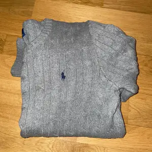 Säljer en snygg grå stickad tröja från Ralph Lauren. Den har ett klassiskt kabelstickat mönster och en liten broderad logga på bröstet. Perfekt för både höst och vinter, och den är i mycket bra skick. Passar till både jeans och chinos!