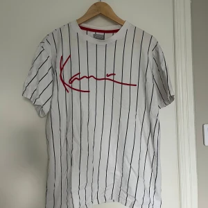 Vit och svart randig t-shirt från Karl Kani - Säljer en snygg vit t-shirt från Karl Kani med svarta ränder och röd logga på bröstet. Perfekt för en avslappnad stil. Den är kortärmad och gjord i mjuk bomull. Passar perfekt till både jeans och shorts!