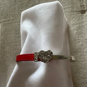 Rött armband med hjärta från Guess - Säljer ett snyggt armband från Guess i rött och silverplätterat stål. Armbandet har ett glittrigt hjärta och små stenar som detaljer. Perfekt för att ge en extra touch till din outfit. Justerbar storlek med en liten kedja och Guess-logga. Passar perfekt till fest eller vardag! Som nytt!!