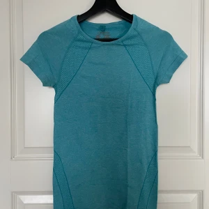 Turkos träningstopp - Säljer en snygg turkos träningstopp i syntetmaterial. Den har korta ärmar och ett coolt mönster som ger en sportig look. Perfekt för gymmet eller löprundan!