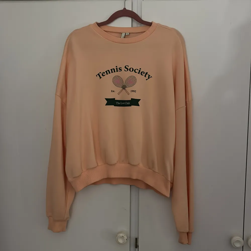 Säljer en superfin persikofärgad sweatshirt från NLY Trend. Den har ett tryck med texten 'Tennis Society' och två tennisrack. Tröjan är i bra skick och perfekt för en avslappnad stil. Den är långärmad och har ribbade muddar vid ärmslut och nederkant. Perfekt för vår och sommar!. Tröjor & Koftor.