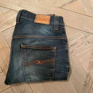 Mörkblå jeans från Nudie Jeans - Säljer ett par snygga mörkblå jeans från Nudie Jeans i bra skick. De har en klassisk femficksdesign med orange sömmar och en cool sliten look. Storlek en är 32W i midjan men är lite små så skulle kunna passa 30/31 och längden är 34 