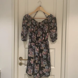 Blommig klänning med trekvartsärm - Säljer en superfin blommig klänning med trekvartsärm. Den har en elastisk offshoulder-design och ett vackert blommönster i rosa, lila och beige på en svart bakgrund. Perfekt för vår och sommar! 🌸