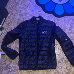 Svart pufferjacka från Emporio Armani - Säljer en snygg svart pufferjacka från Emporio Armani. Jackan har en stilren design med dragkedja framtill och är perfekt för kyligare dagar. Den är i mycket bra skick och passar både till vardags och fest. Stor logga på bröstet ger en exklusiv touch. Men den har ett vitt färg plack så därför sänker ja till 400kr😉