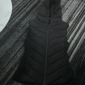 Svart dunväst från Zara - Säljer en snygg svart dunväst från Zara i storlek S. Västen har en praktisk dragkedja och en huva för extra värme. Perfekt för kyliga dagar och kan enkelt matchas med olika outfits. Använd den över en tröja eller jacka för en stilren look.