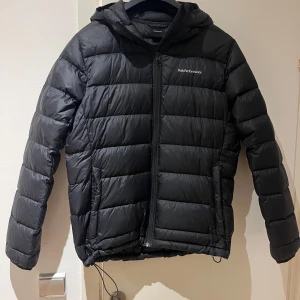 Svart dunjacka från Peak Performance - Säljer en svart dunjacka från Peak Performance i mycket bra skick. Jackan har en stilren design med dragkedja och huva, perfekt för kalla vinterdagar. Den är långärmad och håller dig varm och bekväm. Passar både till vardags och utomhusaktiviteter.