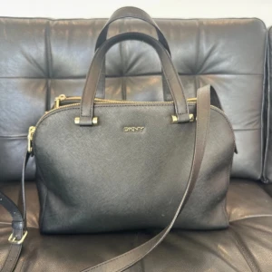 Svart handväska från DKNY - Snygg svart handväska från DKNY i skinn med guldfärgade detaljer och dragkedja. Perfekt för både vardag och fest. Väskan har både handtag och en axelrem för flexibel användning. Den är i mycket bra skick och har en stilren design med DKNY-logga framtill.