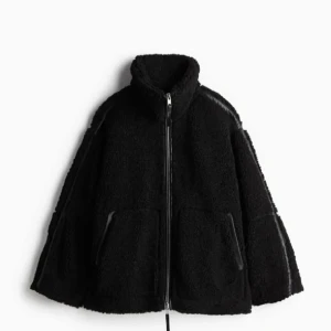 Svart teddyjacka med dragkedja - Mysig svart teddyjacka perfekt för kyliga dagar. Den har en dragkedja framtill och stora fickor på sidorna. Jackan är långärmad och har en hög krage för extra värme. Perfekt för både höst och vinter! Nyskick med lappar kvar