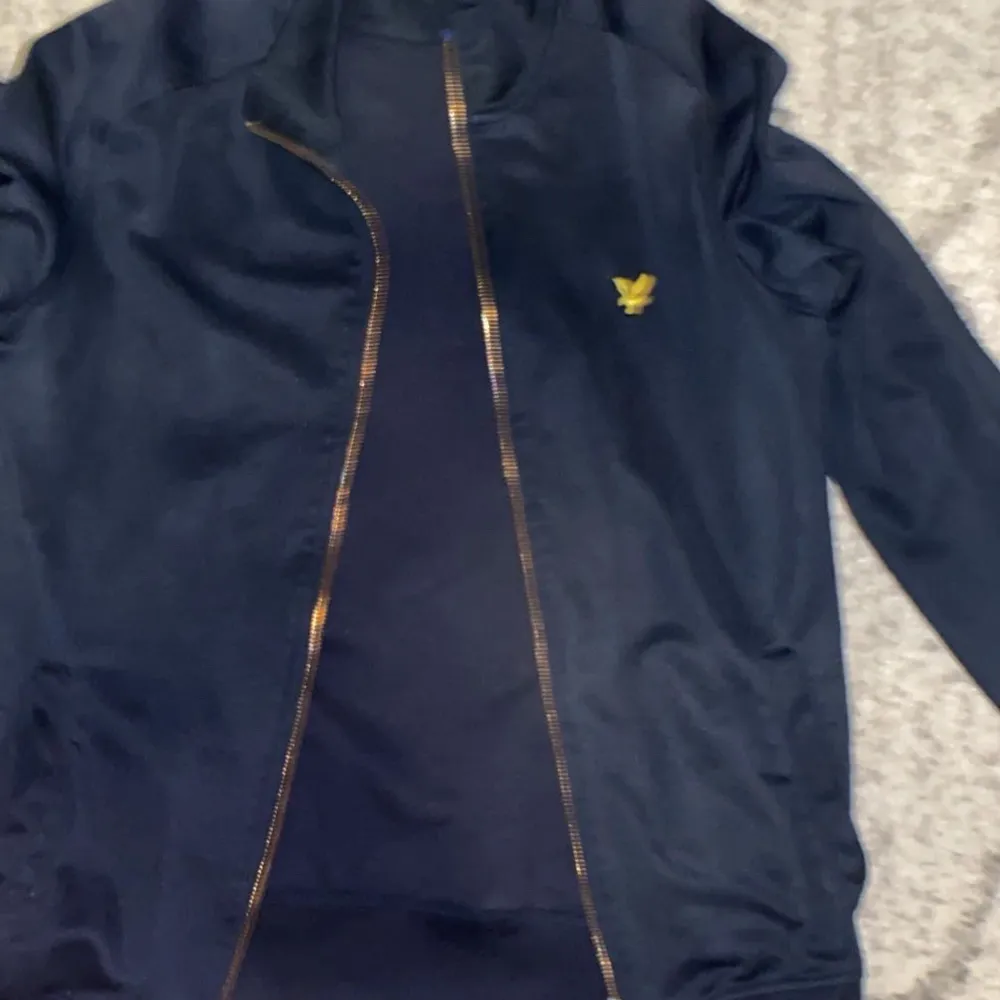 Säljer en snygg mörkblå jacka från Lyle & Scott i mycket bra skick. Den har en dragkedja framtill och ett litet gult logomärke på bröstet. Perfekt för höst och vår, med en stilren design som passar till det mesta. Jackan är i storlek M och har långa ärmar.. Jackor.