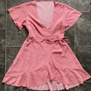 Rosa omlottklänning med prickar - Säljer en superfin rosa omlottklänning med vita prickar. Den har korta ärmar och en söt volangdetalj längst ner. Perfekt för sommarens alla fester! 💖