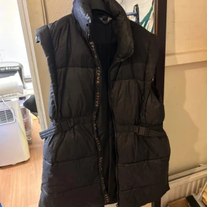 Svart väst med detaljer - Västen är väl använd och är varm och mysig att bära. Den har små detaljer som vid dragkedjan och axeln. Den har även ett band som går att dra åt i midjan