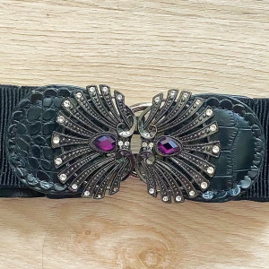 Svart elastiskt bälte med dekorativt spänne - Säljer ett snyggt svart elastiskt bälte med ett riktigt coolt spänne. Spännet har en unik design med lila stenar och silverdetaljer som ger en glamorös touch. Bältet är gjort av ett elastiskt material som gör det bekvämt att bära. Perfekt för att piffa upp vilken outfit som helst!