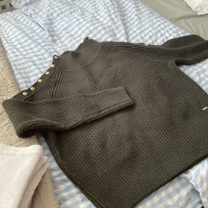 Mörkgrå stickad tröja från Holly & White - Säljer en supermysig mörkgrå stickad tröja från Holly & White. Den har snygga guldknappar längs axeln som ger en extra touch. Perfekt för kyliga höst- och vinterdagar. Tröjan är i bra skick och redo för en ny ägare!