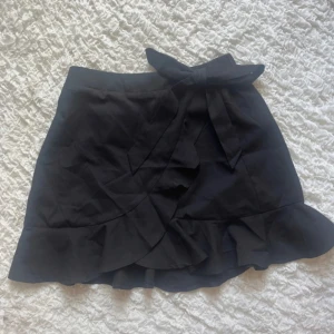 Svart volangkjol - Söt svart kjol. Använd fåtal gånger. Knytningen och omlottdelen är fastsydd alltså ej justerbar. Däremot resår i midjan baktill!