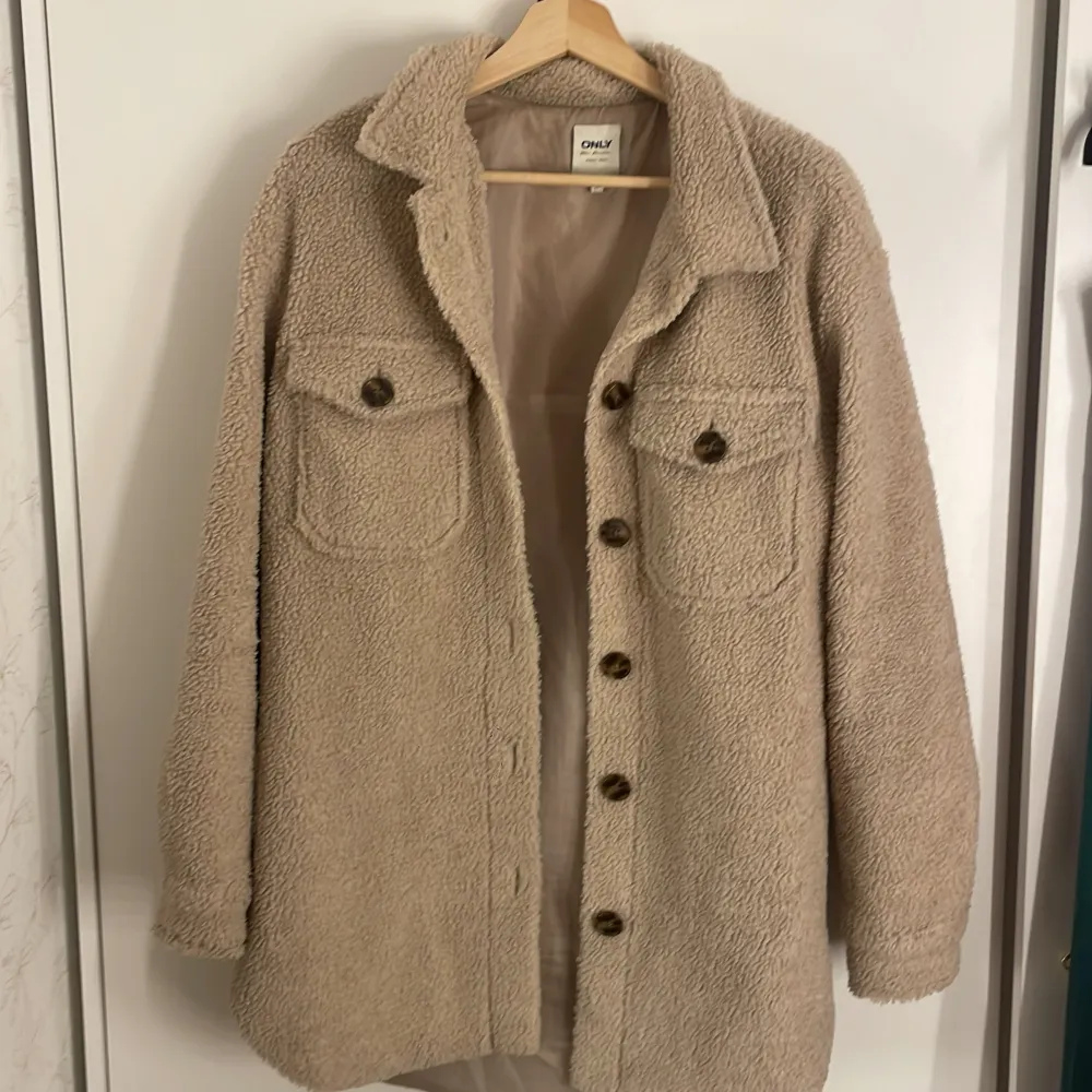 Säljer en supermysig beige teddyjacka från ONLY. Den har en oversized passform och är perfekt för höst och vinter. Jackan har långa ärmar och stängs med knappar framtill. Två stora bröstfickor med knappar ger en cool look. Dock har den lite missfärgning på insida kant, men det är inget märkvärdigt eller något som annars syns.. Jackor.