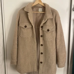 Beige teddyjacka från ONLY - Säljer en supermysig beige teddyjacka från ONLY. Den har en oversized passform och är perfekt för höst och vinter. Jackan har långa ärmar och stängs med knappar framtill. Två stora bröstfickor med knappar ger en cool look. Dock har den lite missfärgning på insida kant, men det är inget märkvärdigt eller något som annars syns.
