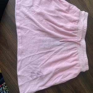 Rosa kortkjol med strassdetaljer - Säljer en superfin rosa kortkjol med elastisk midja. Kjolen har coola strassdetaljer som bildar bokstäver på baksidan och en snygg logga på sidan. Perfekt för en avslappnad men ändå glammig look!