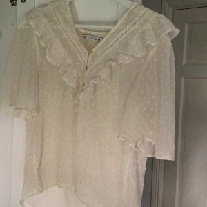 Vit blus från Zara - Säljer en superfin vit blus från Zara i storlek S. Den har korta ärmar och är dekorerad med volanger och spetsdetaljer. Perfekt för sommardagar eller en festlig kväll. Blusen är i nyskick och har en söt knäppning framtill.