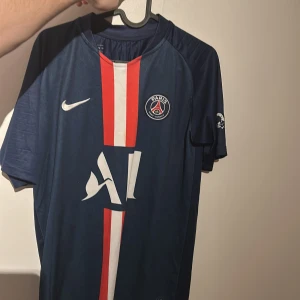 PSG tröja - Snygg blå PSG-tröja från Nike med Mbappé och nummer 7 på ryggen. Tröjan har korta ärmar och ett stilrent mönster med röda och vita detaljer. Perfekt för fotbollsfans som vill visa sitt stöd för laget och spelaren.