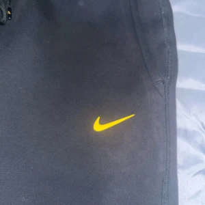Svarta mjukisbyxor från Nike NOCTA - Snygga svarta mjukisbyxor från Nike NOCTA med gul logga. Byxorna har dragkedja och snörning i midjan för en bekväm passform. Perfekta för en avslappnad stil.