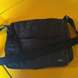 Svart axelväska från Armani Jeans - Snygg svart axelväska från Armani Jeans med subtilt mönster och justerbar axelrem. Väskan har en praktisk ficka framtill och stängs med magnetknapp. Perfekt för att bära med sig det nödvändigaste med stil.