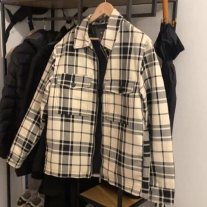 Rutig overshirt i beige och svart - Säljer en snygg rutig overshirt i beige och svart med dragkedja framtill. Jackan har långa ärmar och två stora bröstfickor med knappar. Perfekt för lager-på-lager-stilen!