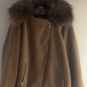 Brun kappa med fuskpäls - Snygg brun kappa med fuskpäls vid kragen och dragkedja framtill. Perfekt för kyliga dagar med sin stilrena design och praktiska fickor med dragkedjor.