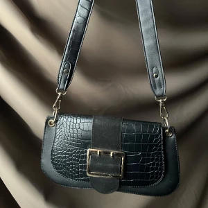 Svart axelväska med krokodilmönster och spänne - Snygg svart axelväska med krokodilmönster och en stor dekorativ spänne framtill. Väskan har en avtagbar axelrem och är perfekt för att ge en stilren touch till din outfit.