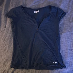 Mörkblå kortärmad topp från Hollister - Säljer en mörkblå topp från Hollister med korta ärmar och bruna knappar framtill. En liten logga vid nederkanten. Kort