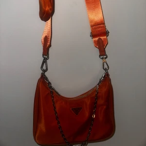 Orange axelväska med kedjedetalj - Snygg orange axelväska med justerbar axelrem och kedjedetalj. Väskan har en dragkedja och en liten matchande väska fäst vid remmen. Perfekt för att ge en färgklick till din outfit.