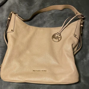 Beige axelväska från Michael Kors - Säljer en stilren beige axelväska från Michael Kors i skinn. Väskan har en praktisk dragkedja och ett elegant hängsmycke med MK-logga. Perfekt för att bära dina dagliga nödvändigheter med stil.